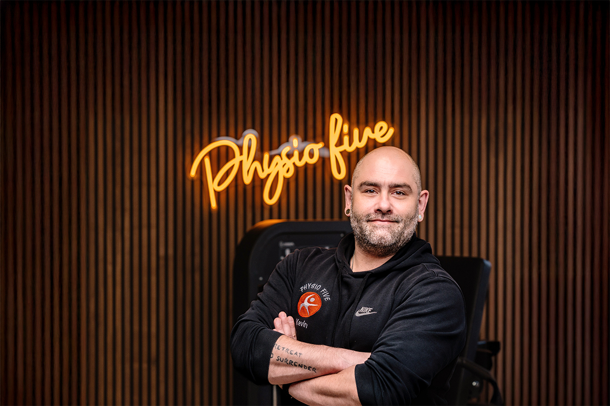 Ein Mitarbeiter von Physio Five, einer modernen Physiotherapiepraxis mit angeschlossenem Gesundheitsstudio in Merenberg, steht mit gekreuzten Armen vor einem neonleuchtenden 