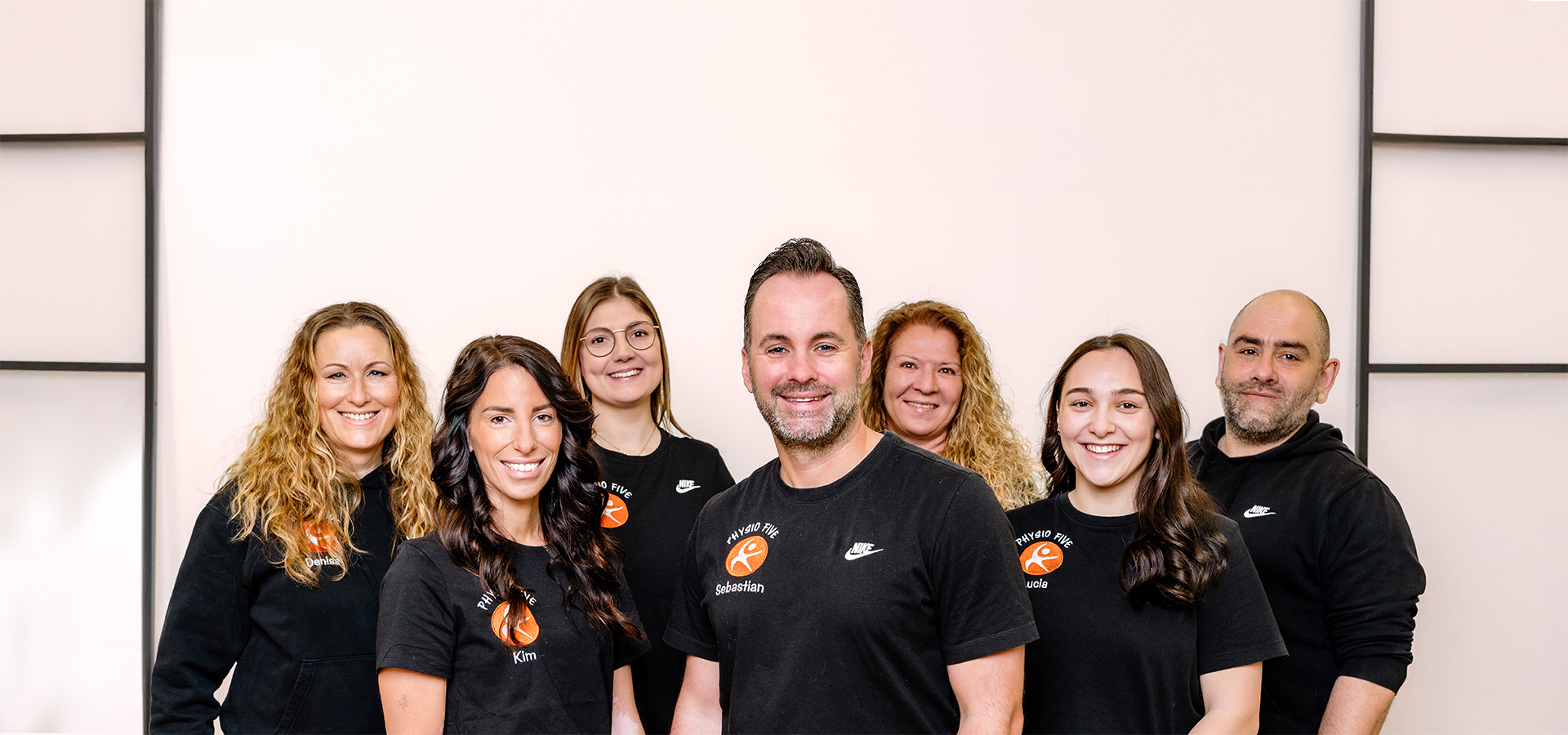 Das Team der modernen Physiotherapiepraxis Physio Five mit angeschlossenem Gesundheitsstudio in Merenberg posiert für ein Gruppenfoto. Die Mitglieder tragen schwarze Shirts mit dem Praxislogo und Namen wie Sebastian, Kim oder Lucila.