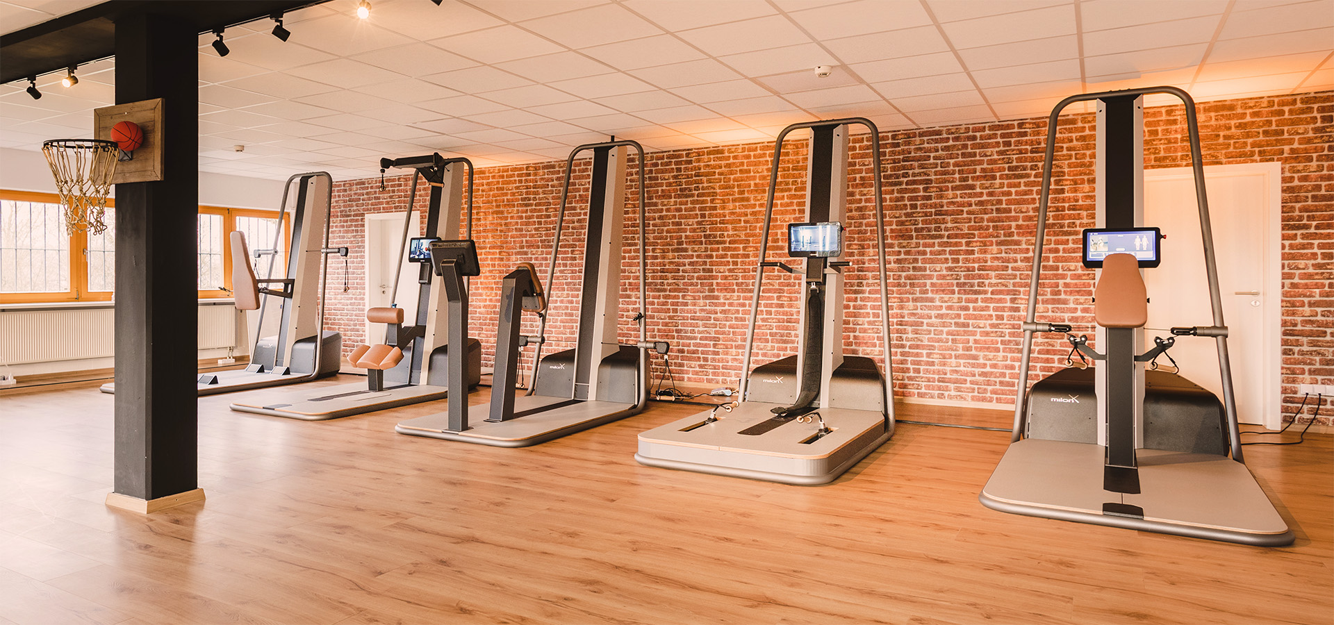 Modernes Gesundheitsstudio von Physio Five mit milon- und milon X-Trainingsgeräten, Backsteinwand und Holzfußboden – Raum für medizinisches Gesundheitstraining, Physiotherapie und individuelle Betreuung.