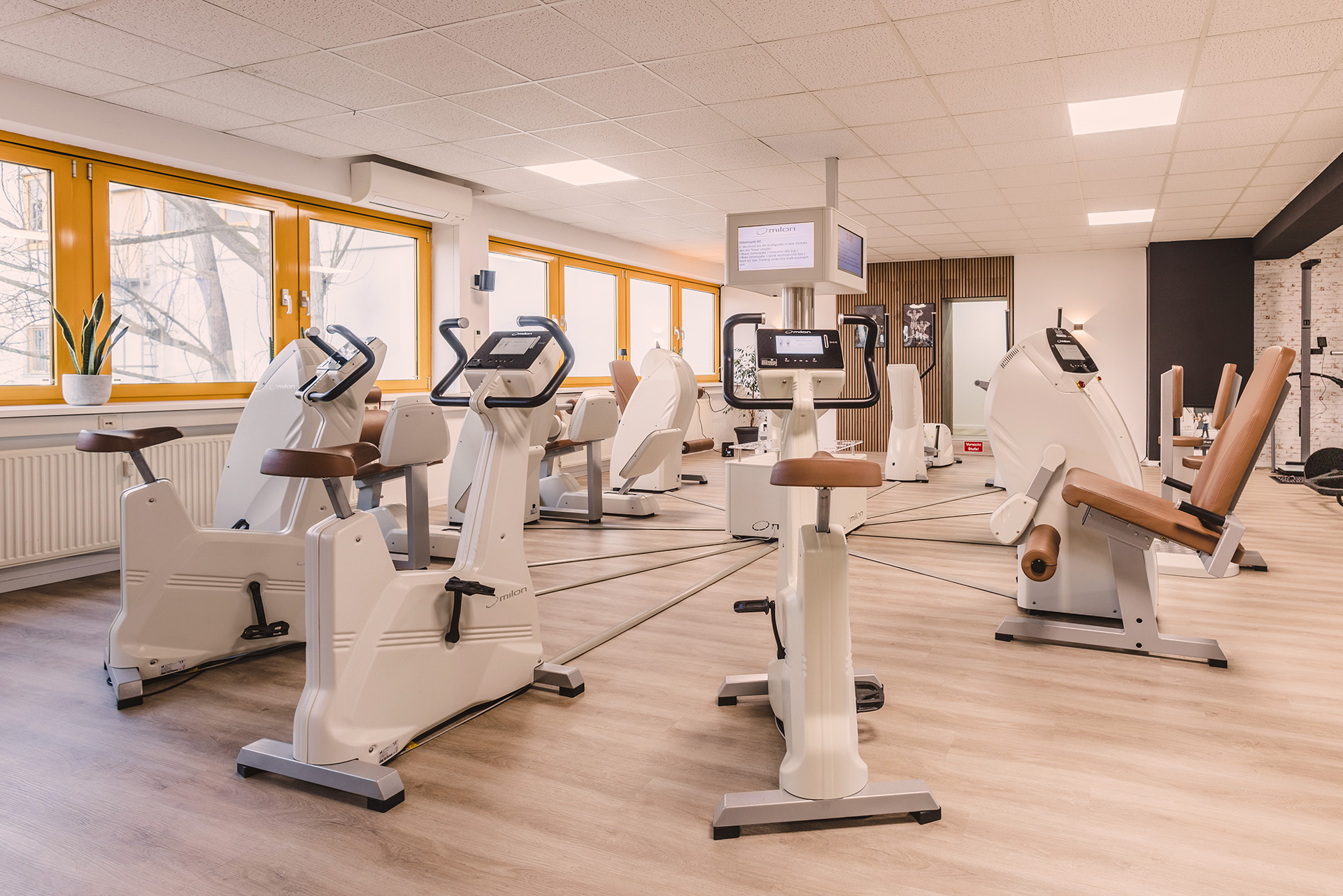 Modernes Gesundheitsstudio der Physio Five Praxis mit milon-Trainingsgeräten, hellen Fenstern und hellen Holzböden – Raum für medizinisches Gesundheitstraining und Physiotherapie.