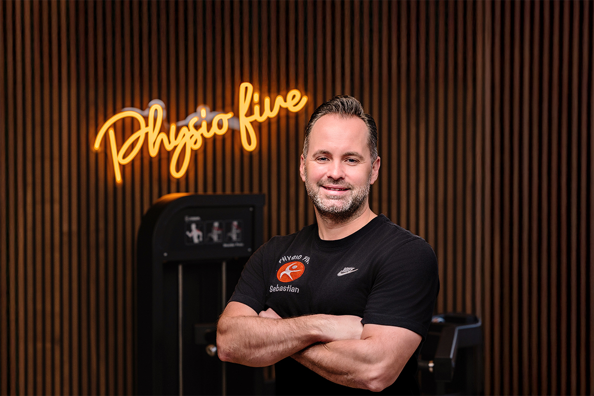 Ein Mann in einem schwarzen Physio Five Shirt steht vor einem neonbeleuchteten 