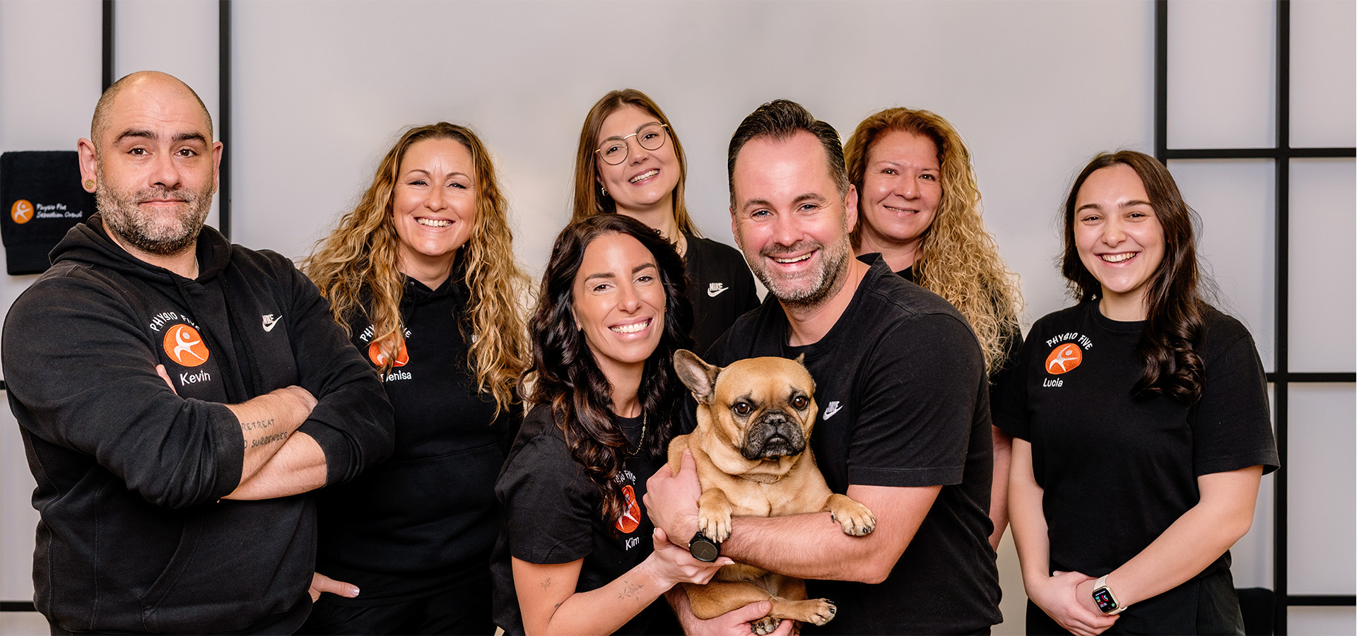 Das Team von Physio Five, einer modernen Physiotherapiepraxis mit angeschlossenem Gesundheitsstudio in Merenberg, posiert gemeinsam in brandeden Shirts – mit einem freundlichen Französischen Bulldoggen, der die herzliche Atmosphäre unterstreicht.