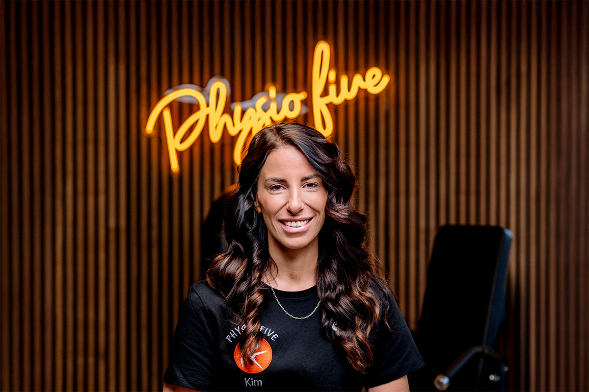 Kim, Mitarbeiterin der modernen Physiotherapiepraxis Physio Five in Merenberg, steht vor dem neonbeleuchteten Praxislogo, das die Kombination aus Physiotherapie und Gesundheitstraining repräsentiert.