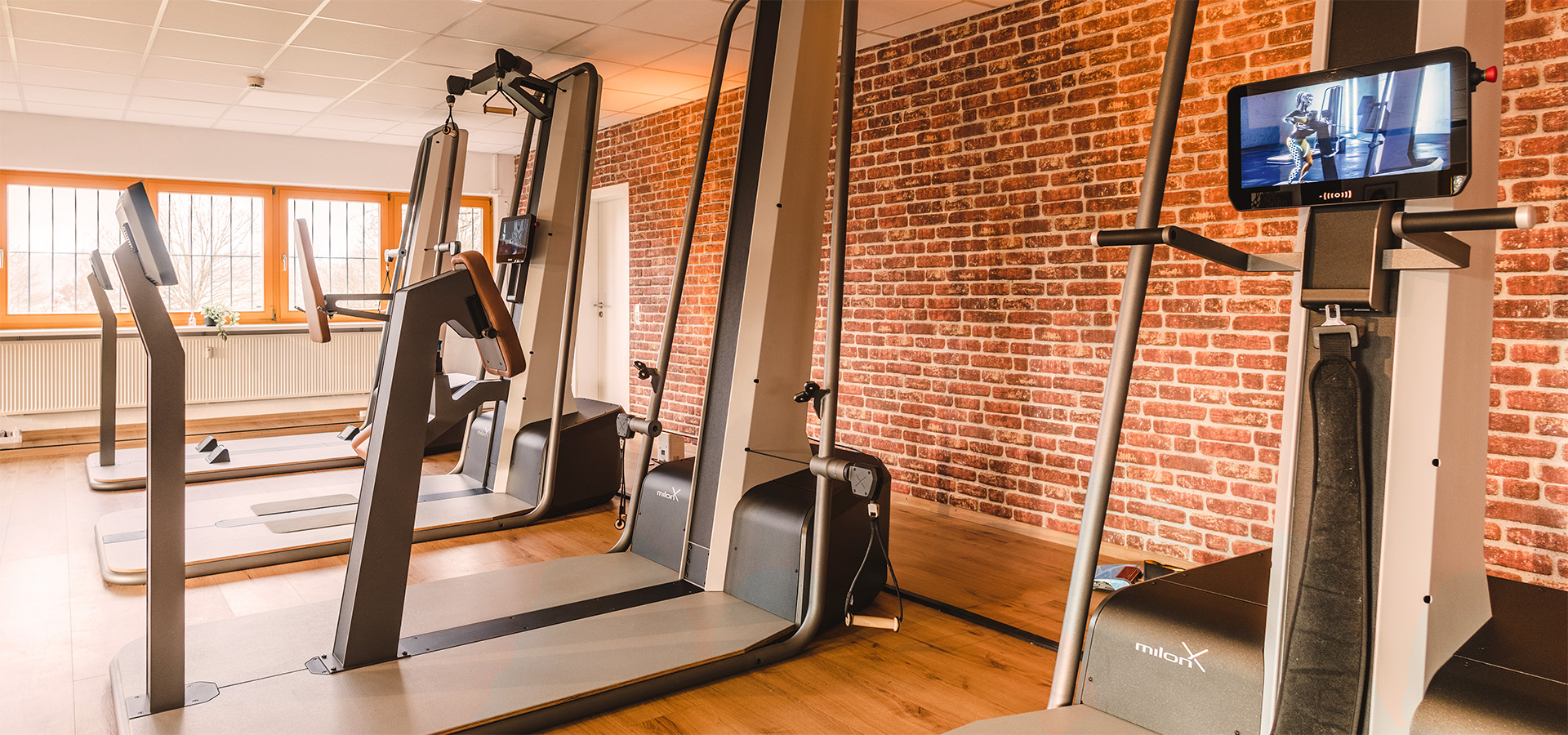 Modernes Fitnessstudio der Physio Five Praxis mit milon- und milon X-Trainingsgeräten, Ziegelwand und großen Fenstern für medizinisches Gesundheitstraining.
