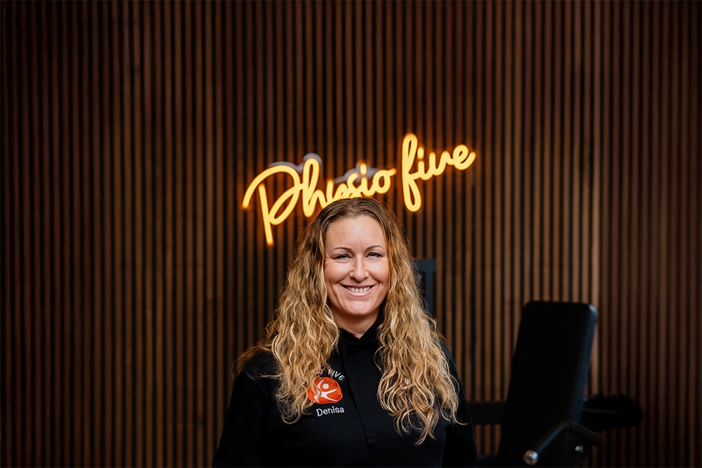 Denisa, Mitarbeiterin von Physio Five, einer modernen Physiotherapiepraxis mit angeschlossenem Gesundheitsstudio in Merenberg, steht vor einem holzvertäfelten Wandelement mit dem neonbeleuchteten Logo 